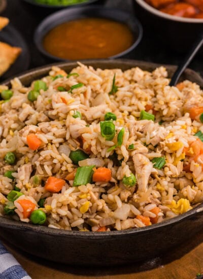 Chicken fried rice in a skillet, garnished with green onions, surrounded by egg rolls and sweet sauces.