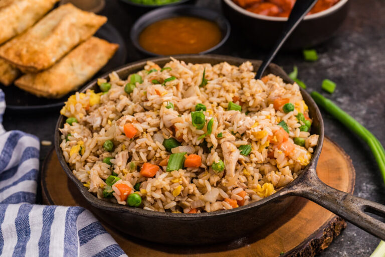 Chicken fried rice in a skillet, garnished with green onions, surrounded by egg rolls and sweet sauces.