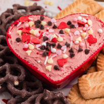 Heart-shaped bowl of Valentine’s cheesecake dip with sprinkles, paired with pretzels and rectangular cookies on a dessert platter.