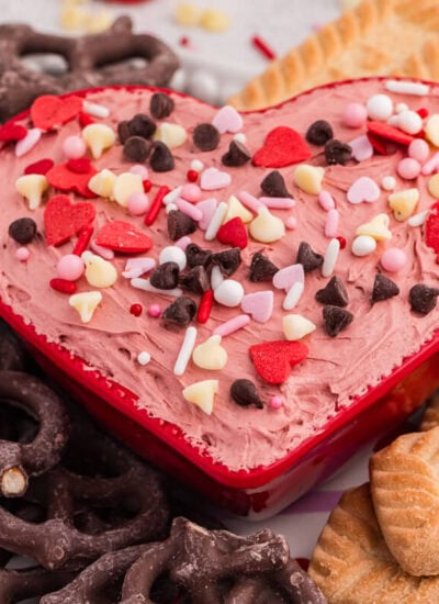 Heart-shaped bowl of Valentine&rsquo;s cheesecake dip with sprinkles, paired with pretzels and rectangular cookies on a dessert platter.