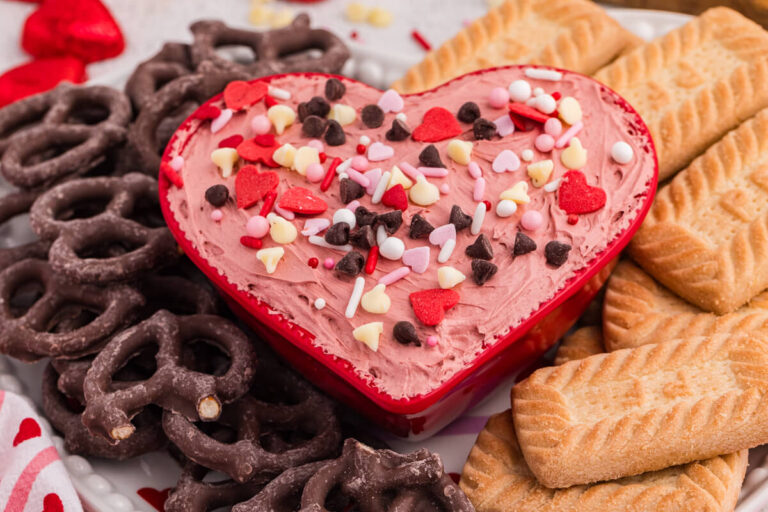 Heart-shaped bowl of Valentine’s cheesecake dip with sprinkles, paired with pretzels and rectangular cookies on a dessert platter.