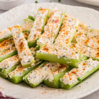 Plate of stuffed celery sticks topped with paprika seasoning and creamy cheese filling, styled with olives, carrot sticks, and fresh chives on a bright kitchen surface.