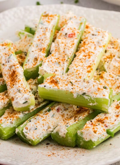 Plate of stuffed celery sticks topped with paprika seasoning and creamy cheese filling, styled with olives, carrot sticks, and fresh chives on a bright kitchen surface.