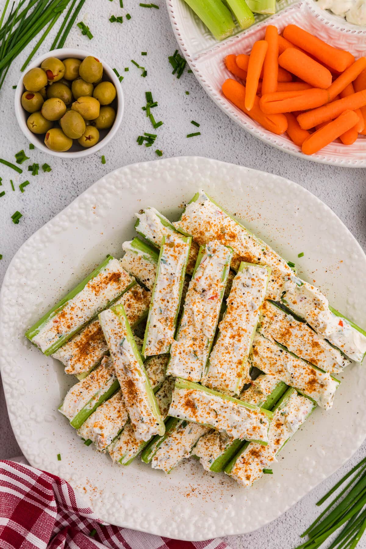 Celery sticks stuffed with a creamy herb cheese mixture and topped with paprika and black pepper, plated on a white dish with olives, carrots, and chopped chives in the background.