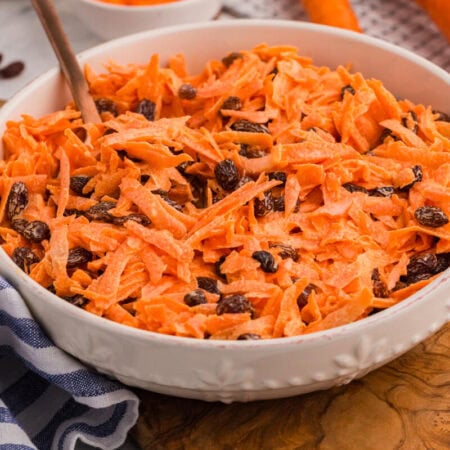 Close-up of creamy shredded carrot salad with raisins in a white ceramic bowl, with fresh carrots and grated carrot prep blurred in the background.