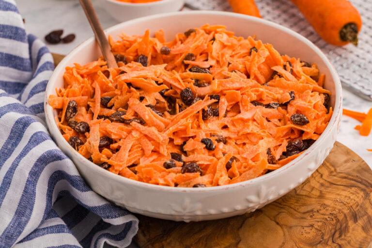 Close-up of creamy shredded carrot salad with raisins in a white ceramic bowl, with fresh carrots and grated carrot prep blurred in the background.