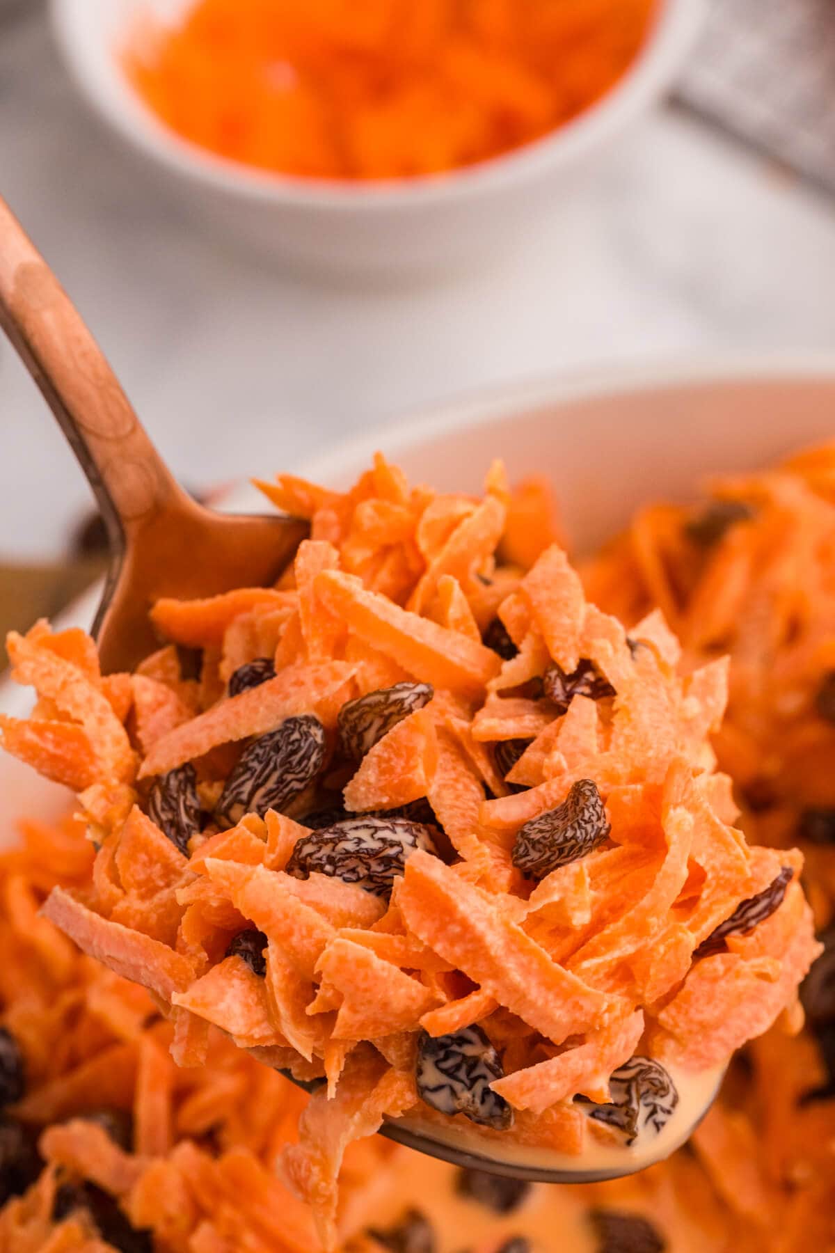 Spoon lifting a serving of shredded carrot and raisin salad, showing detailed texture of carrots and raisins with soft background blur.