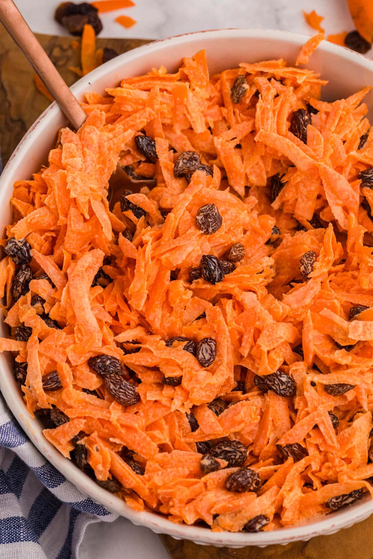 Overhead shot of shredded carrot and raisin salad in a bowl, styled on a wooden board with a striped cloth and scattered carrot shavings.