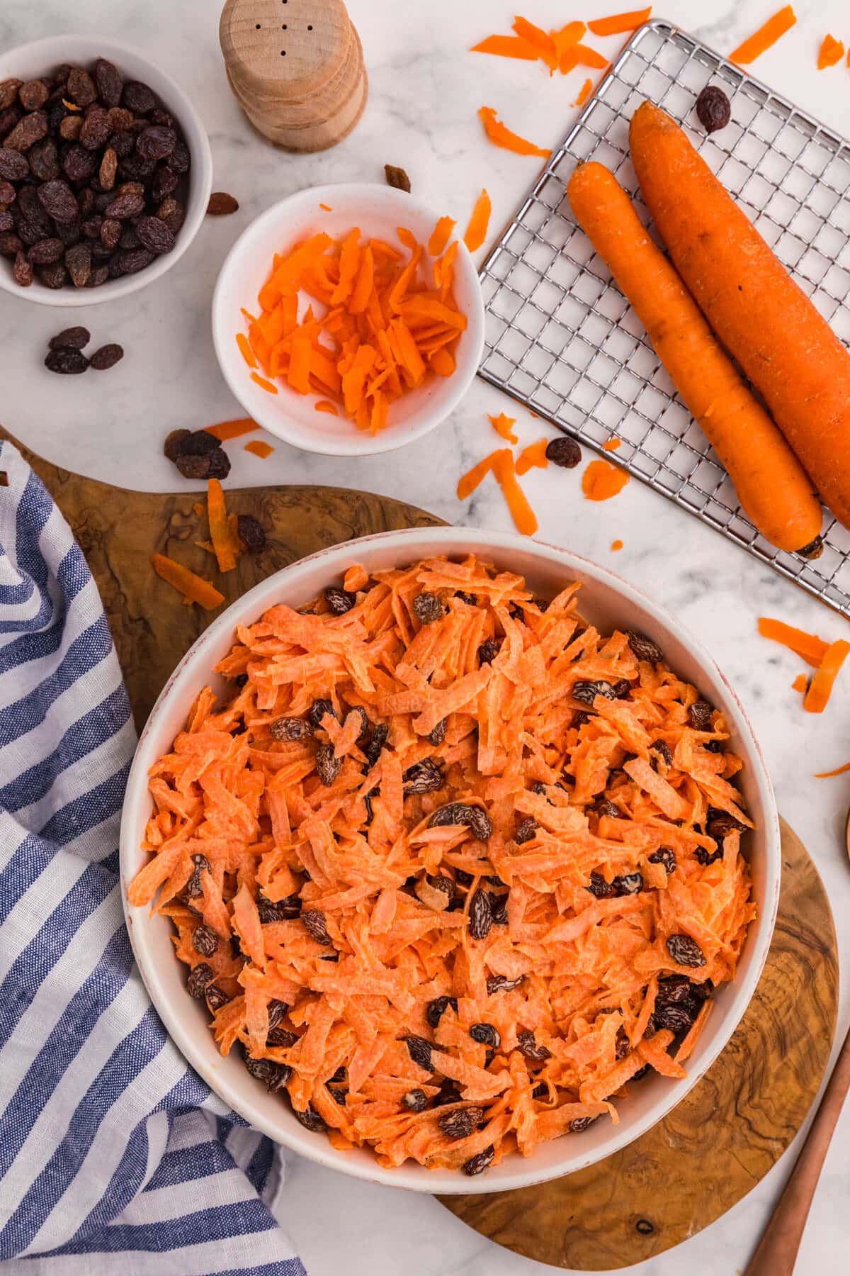 Top-down view of creamy shredded carrot raisin salad in a white bowl, surrounded by whole carrots, grated carrots, a bowl of raisins, and a marble countertop.
