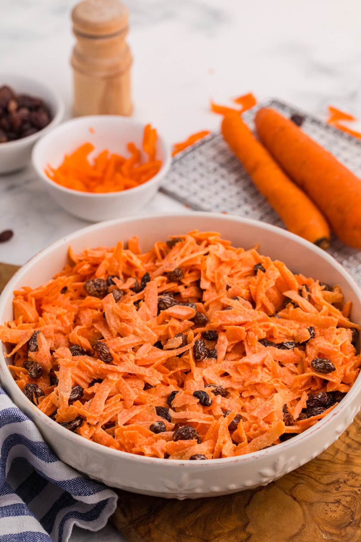 Close-up of creamy shredded carrot salad with raisins in a white ceramic bowl, with fresh carrots and grated carrot prep blurred in the background.