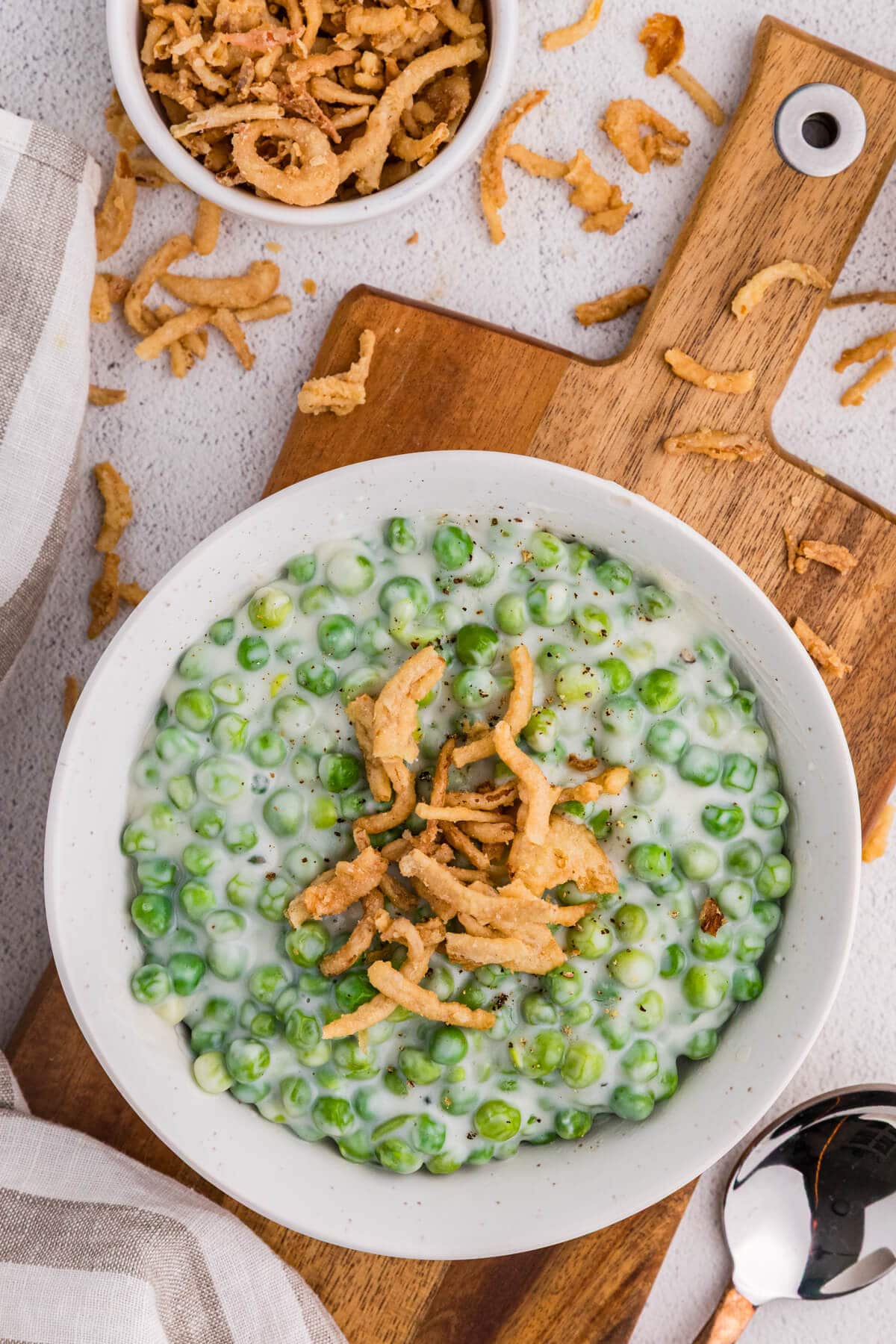 Creamed peas in a white bowl topped with crispy fried onions on a wooden cutting board, overhead view with scattered onions.
