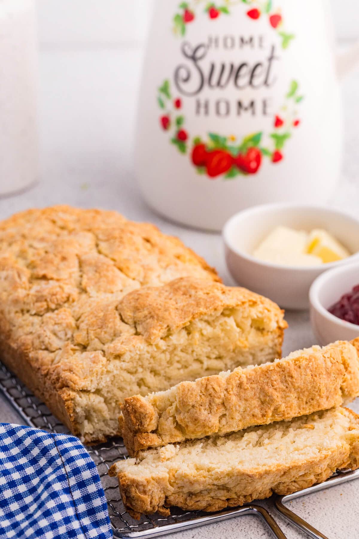 Homemade biscuit bread loaf sliced on a cooling rack with butter and jam, &ldquo;Home Sweet Home&rdquo; jar in background.