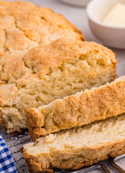 Sliced biscuit bread on wire rack with golden crust, served with butter, jam, and blue gingham cloth.