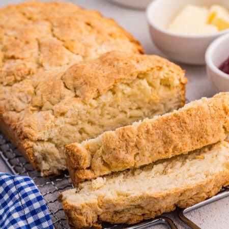 Sliced biscuit bread on wire rack with golden crust, served with butter, jam, and blue gingham cloth.