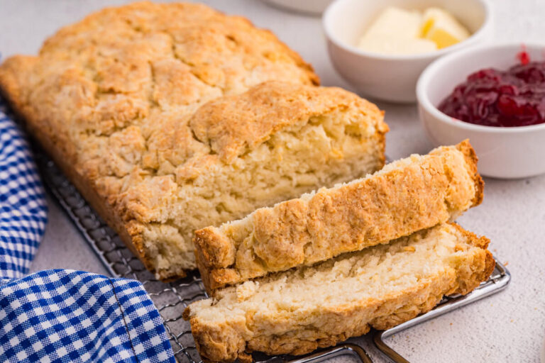 Sliced biscuit bread on wire rack with golden crust, served with butter, jam, and blue gingham cloth.