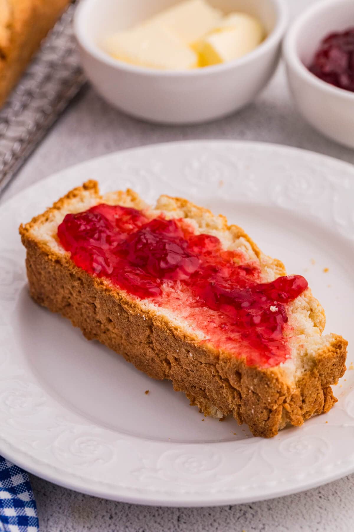 Slice of biscuit bread with red berry jam on white plate, soft interior and crumbly crust.