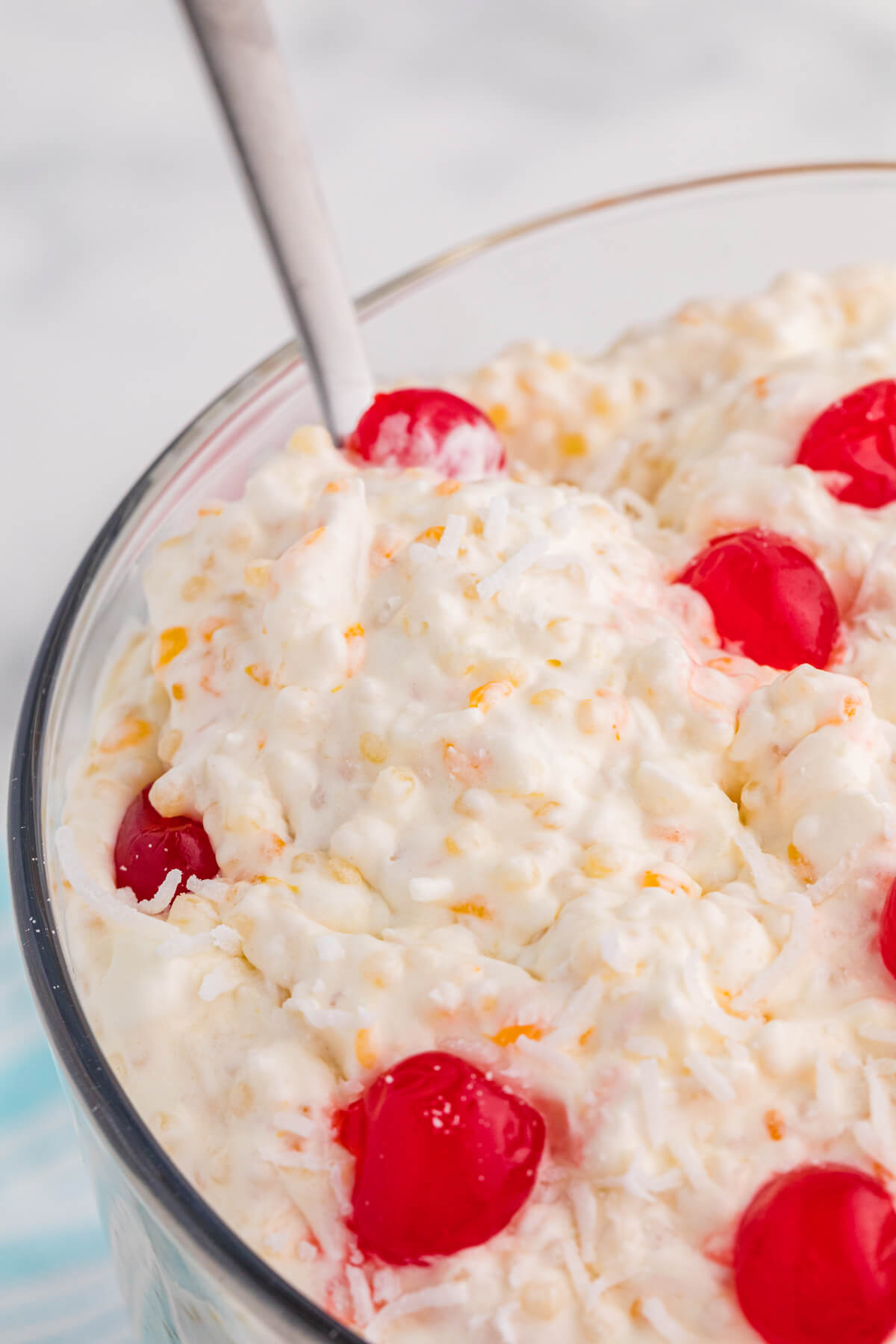 Close-up of a thick, creamy dessert mixture with small pasta pearls, coconut shreds, fruit bits, and glossy red cherries with a spoon inserted.