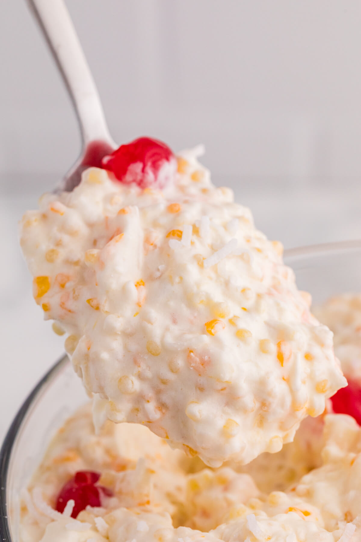 Spoonful of rich, creamy dessert showing soft pasta pearls, coconut flakes, and fruit pieces lifted above the bowl with a cherry on top.