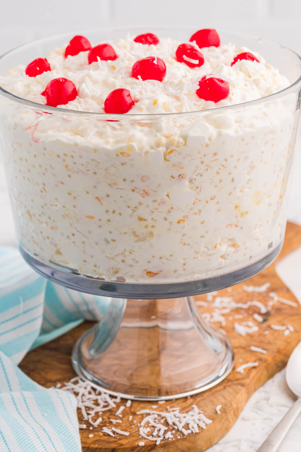 Frog Eye Salad in a clear glass trifle bowl topped with whipped cream, shredded coconut, and maraschino cherries on a wooden board.