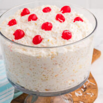 Creamy dessert salad in a clear glass bowl topped with whipped topping, shredded coconut flakes, and bright red maraschino cherries on a wooden serving board.