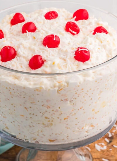 Creamy dessert salad in a clear glass bowl topped with whipped topping, shredded coconut flakes, and bright red maraschino cherries on a wooden serving board.