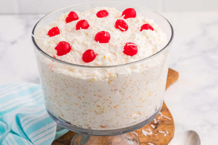 Creamy dessert salad in a clear glass bowl topped with whipped topping, shredded coconut flakes, and bright red maraschino cherries on a wooden serving board.