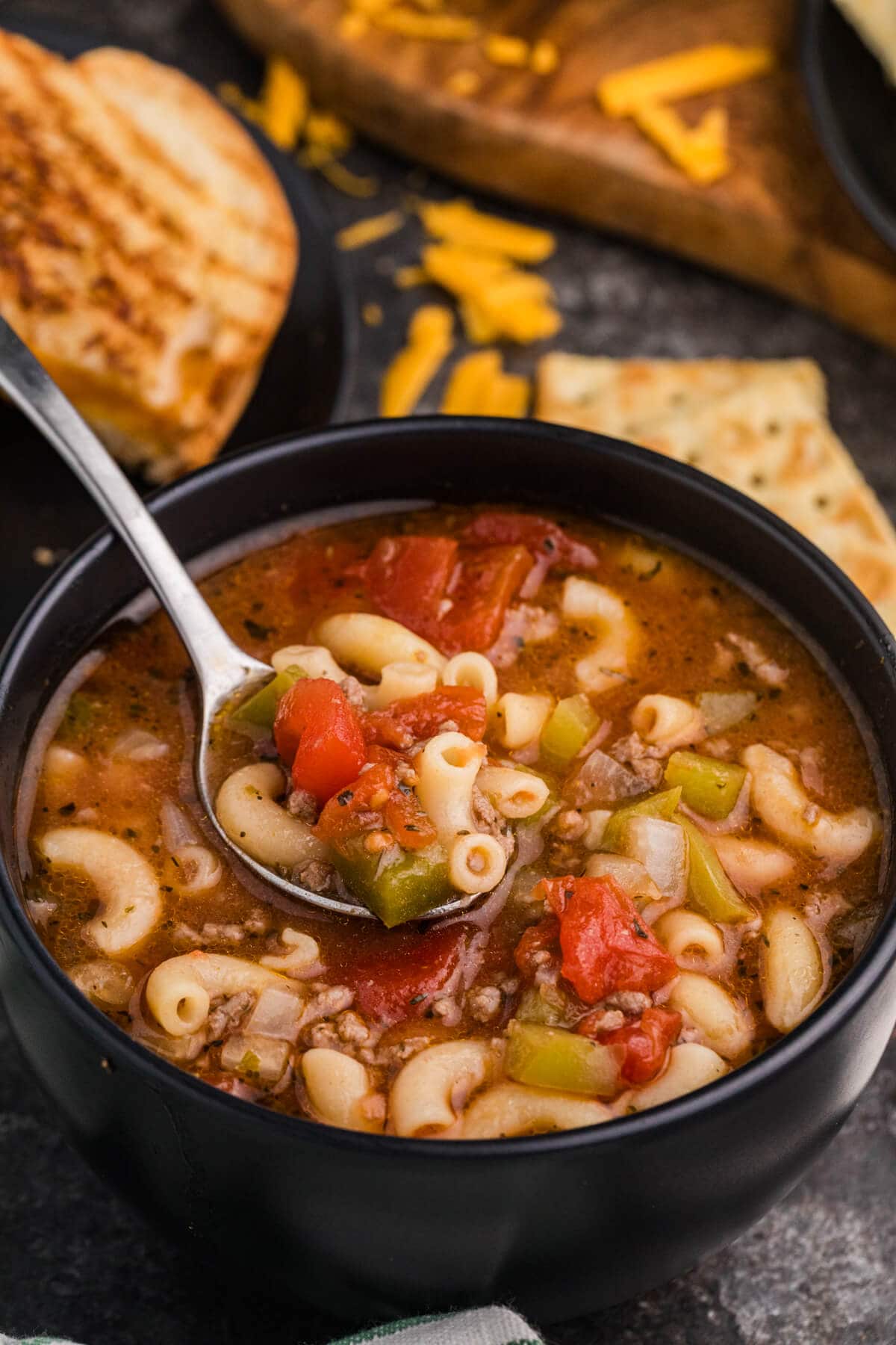 Spoon lifting chunky beef and macaroni tomato soup with elbow pasta, diced tomatoes, onions, and green peppers from a black bowl, with grilled cheese and crackers in the background.