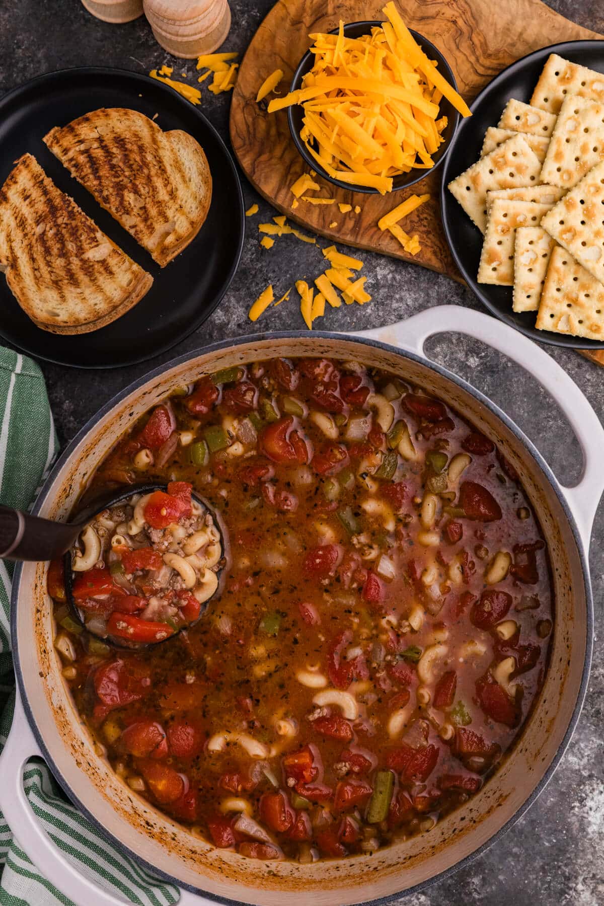 Goulash Soup in a large white Dutch oven filled with tomato-based beef and macaroni soup with diced tomatoes, onions, and green peppers, served alongside shredded cheddar cheese, crackers, and grilled cheese sandwiches on a rustic kitchen surface.