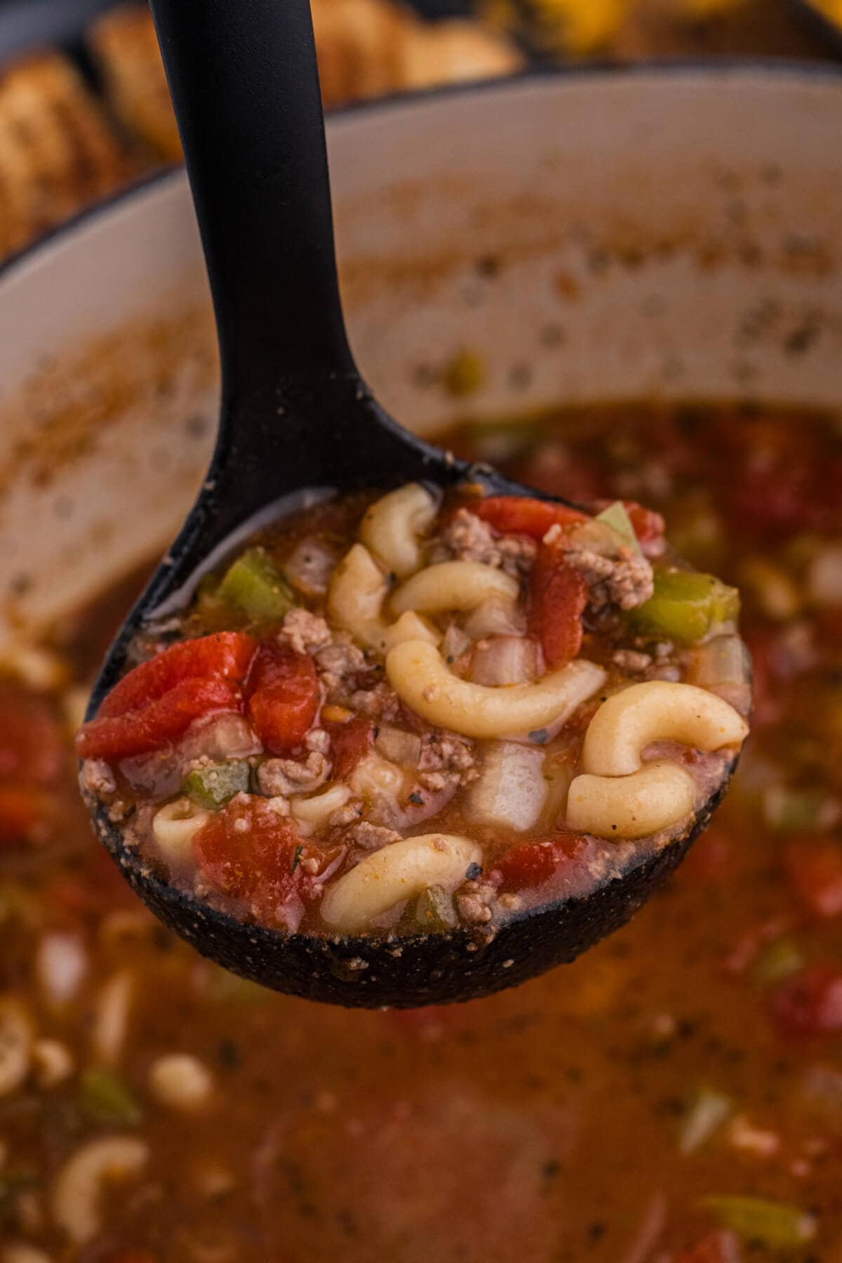 Close-up of a black ladle lifting hearty beef and macaroni soup with elbow pasta, ground beef, diced tomatoes, onions, and green bell peppers in a rich tomato broth.