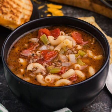 Black bowl filled with beef and macaroni vegetable soup featuring elbow pasta, ground beef, tomatoes, onions, and green peppers, served with grilled cheese sandwiches and crackers on a dark tabletop.