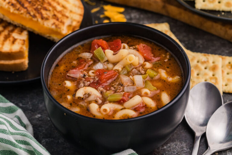 Black bowl filled with beef and macaroni vegetable soup featuring elbow pasta, ground beef, tomatoes, onions, and green peppers, served with grilled cheese sandwiches and crackers on a dark tabletop.