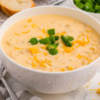 Cheesy elbow pasta soup garnished with green onions and melted cheddar in white bowl, served with bread slices and spoons on marble surface, warm comfort dish.