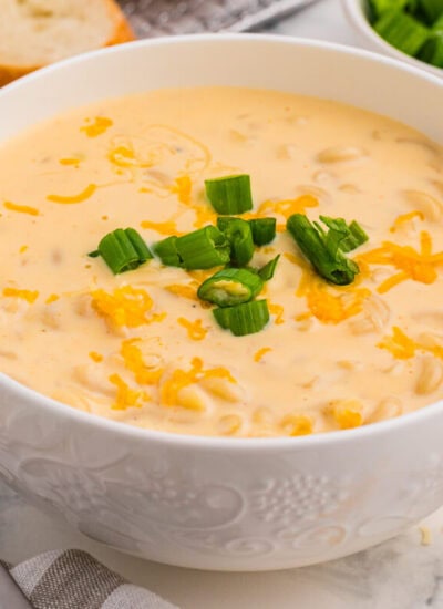 Cheesy elbow pasta soup garnished with green onions and melted cheddar in white bowl, served with bread slices and spoons on marble surface, warm comfort dish.
