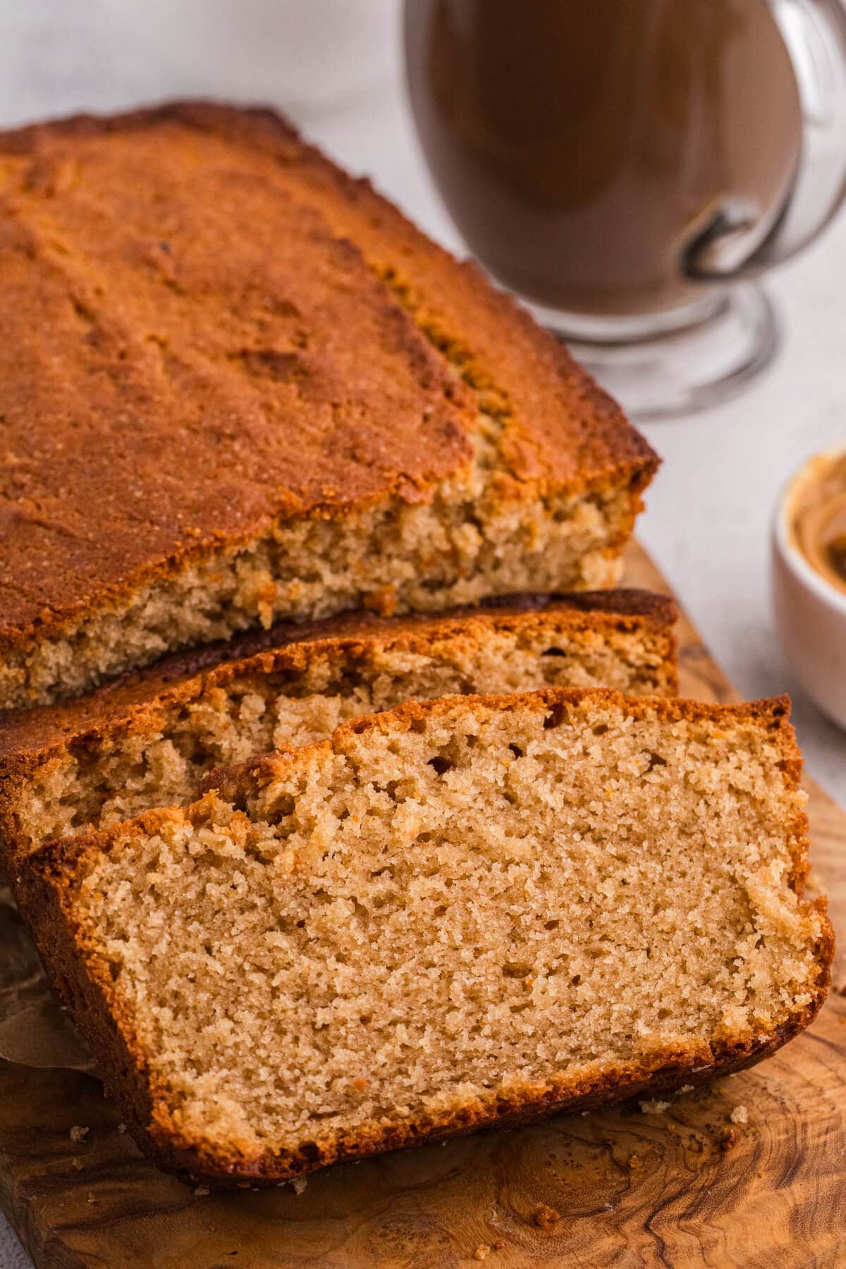 Close-up of sliced peanut butter bread on a wooden cutting board, highlighting moist texture and golden crust with coffee blurred behind.