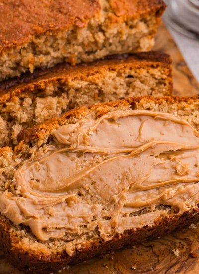Thick slices of peanut butter bread spread with creamy peanut butter on a rustic wooden board, with a small bowl of peanut butter nearby.