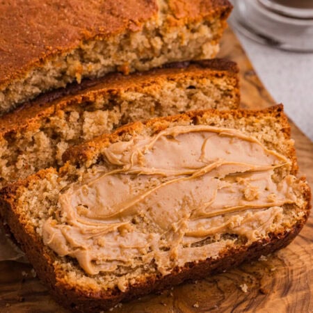 Thick slices of peanut butter bread spread with creamy peanut butter on a rustic wooden board, with a small bowl of peanut butter nearby.