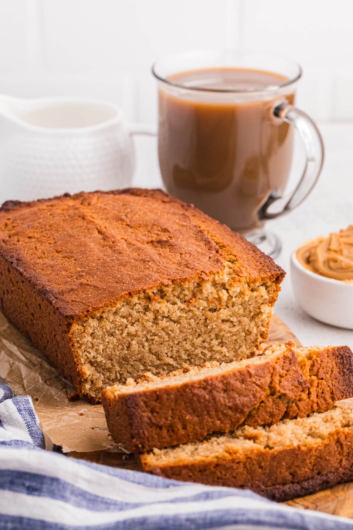 Freshly baked peanut butter bread loaf on parchment paper, sliced to reveal soft crumb, with a glass mug of coffee and a bowl of peanut butter in the background.