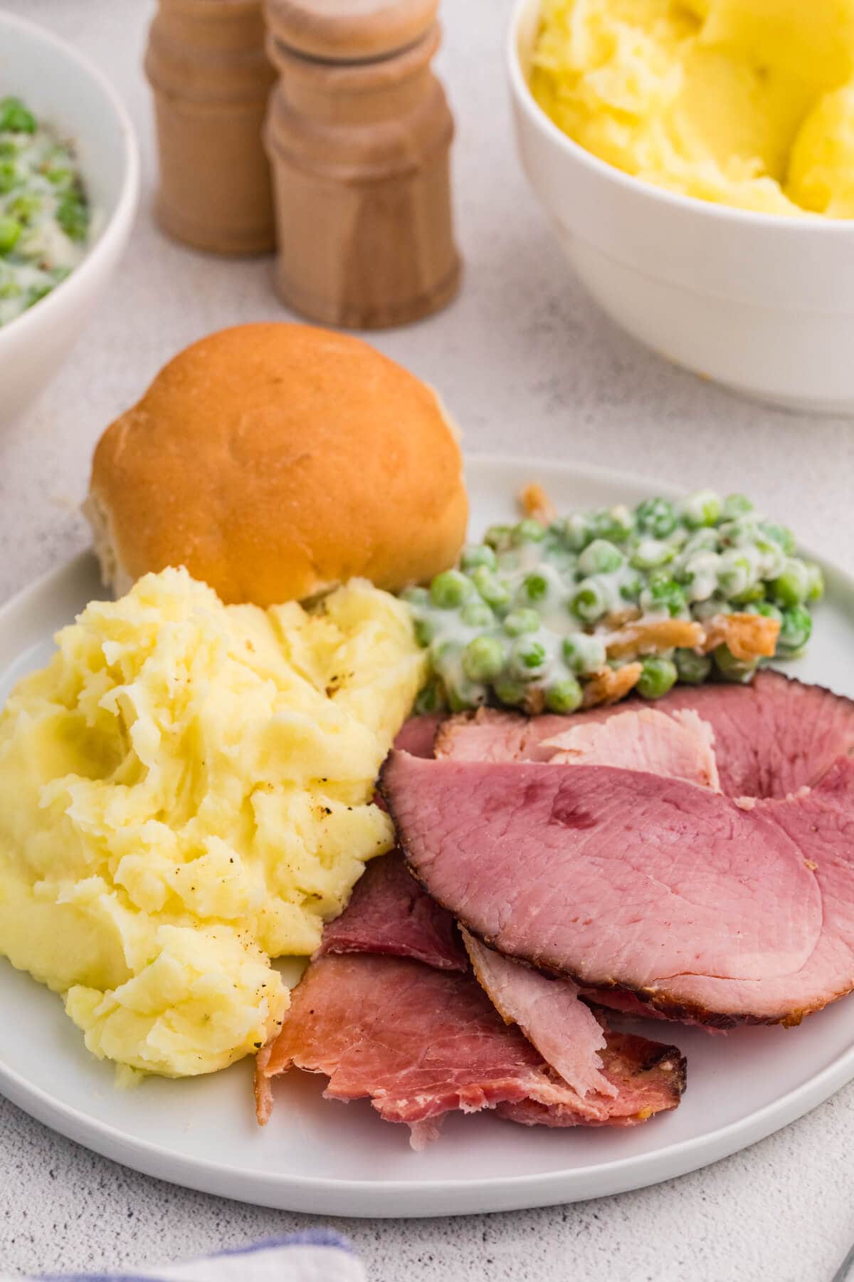 Ham dinner plate with mashed potatoes, creamy peas, and dinner roll, classic comfort food holiday meal setting.