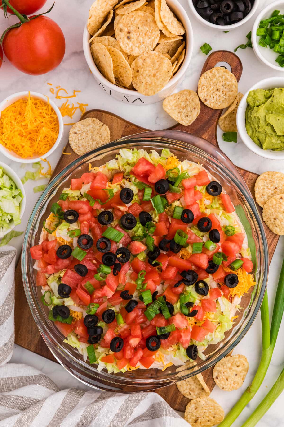 Taco Dip layered appetizer in glass dish topped with diced tomatoes, black olives, green onions, shredded lettuce and cheddar cheese, surrounded by tortilla chips, guacamole, salsa, and fresh ingredients on marble countertop.