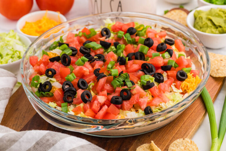 Macro shot of tortilla chip dipped in taco dip with visible layers of sour cream, salsa, shredded cheese, lettuce, and green onions, highlighting creamy texture and fresh toppings.