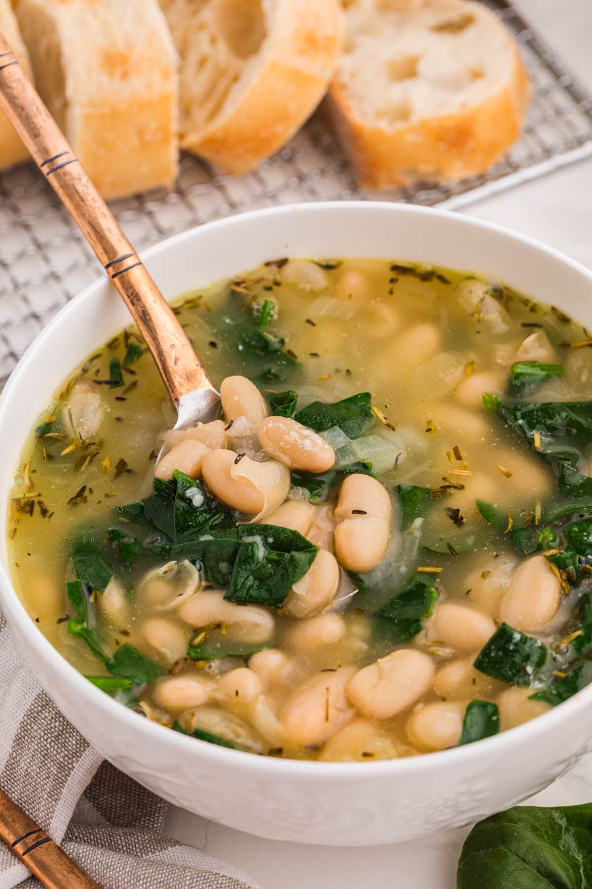 Spoon lifting cannellini beans and spinach from a bowl of Tuscan white bean soup, with crusty bread behind.