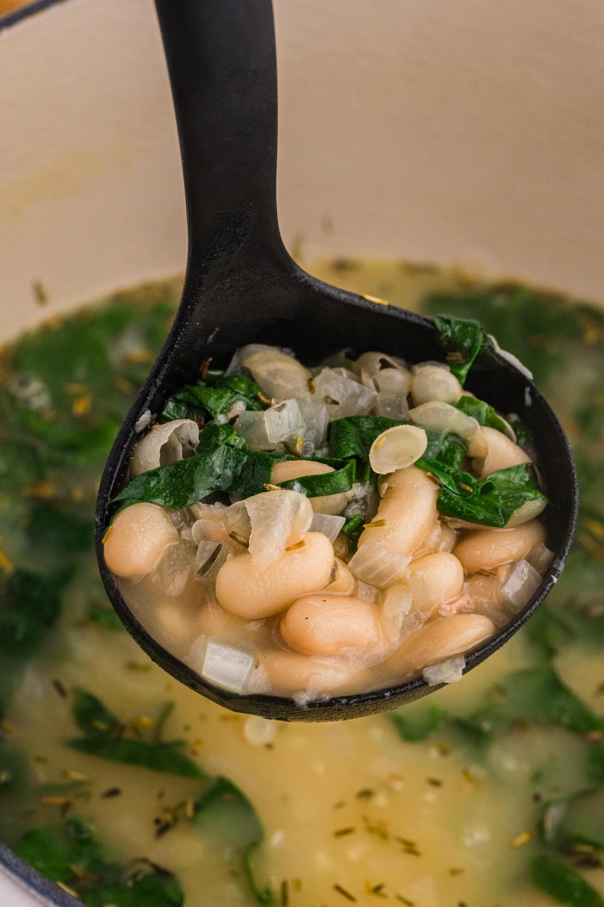 Close-up of white bean soup with cannellini beans, spinach, diced onions, and herbs in broth, served on a black ladle.