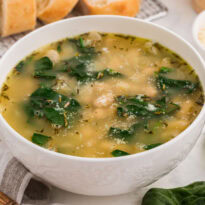 White bowl of Italian white bean and spinach soup with herbs and parmesan, styled with bread and fresh greens on a table.