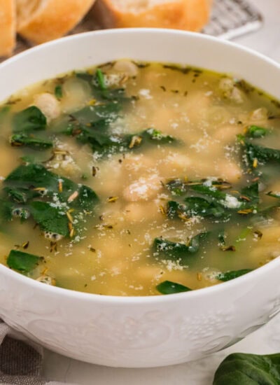 White bowl of Italian white bean and spinach soup with herbs and parmesan, styled with bread and fresh greens on a table.