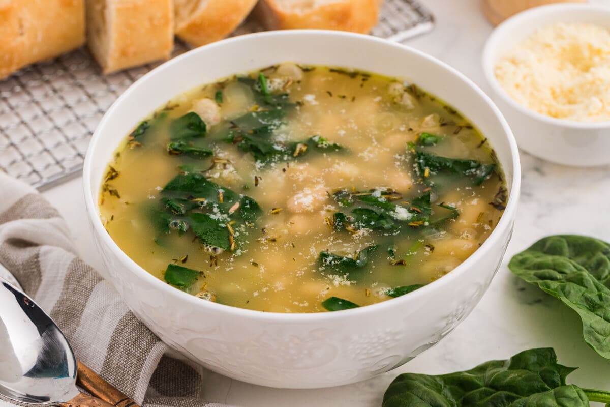 White bowl of Italian white bean and spinach soup with herbs and parmesan, styled with bread and fresh greens on a table.