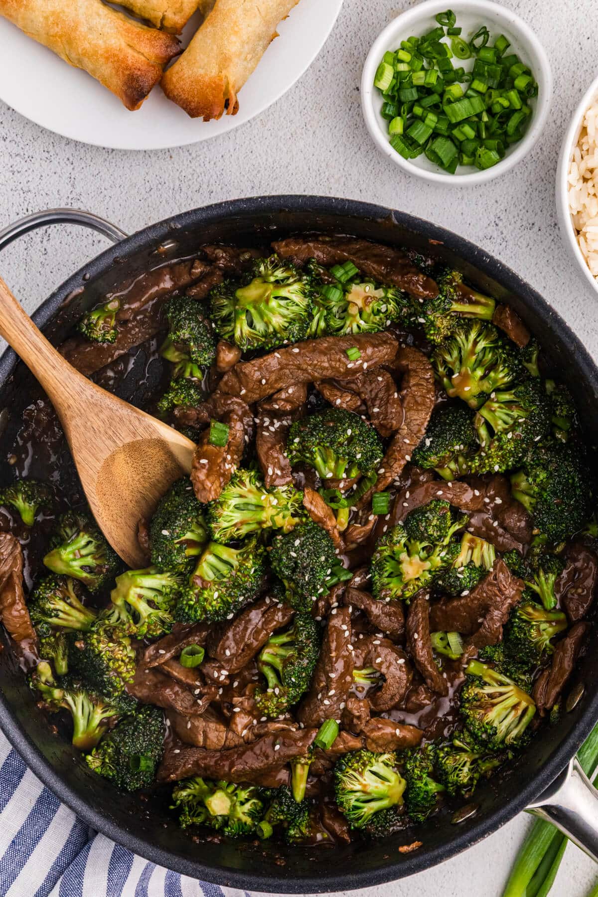 Overhead view of beef and broccoli stir fry in a black skillet with glossy brown sauce, sesame seeds, and chopped green onions, served alongside spring rolls, white rice, and a small bowl of sliced scallions on a light countertop.