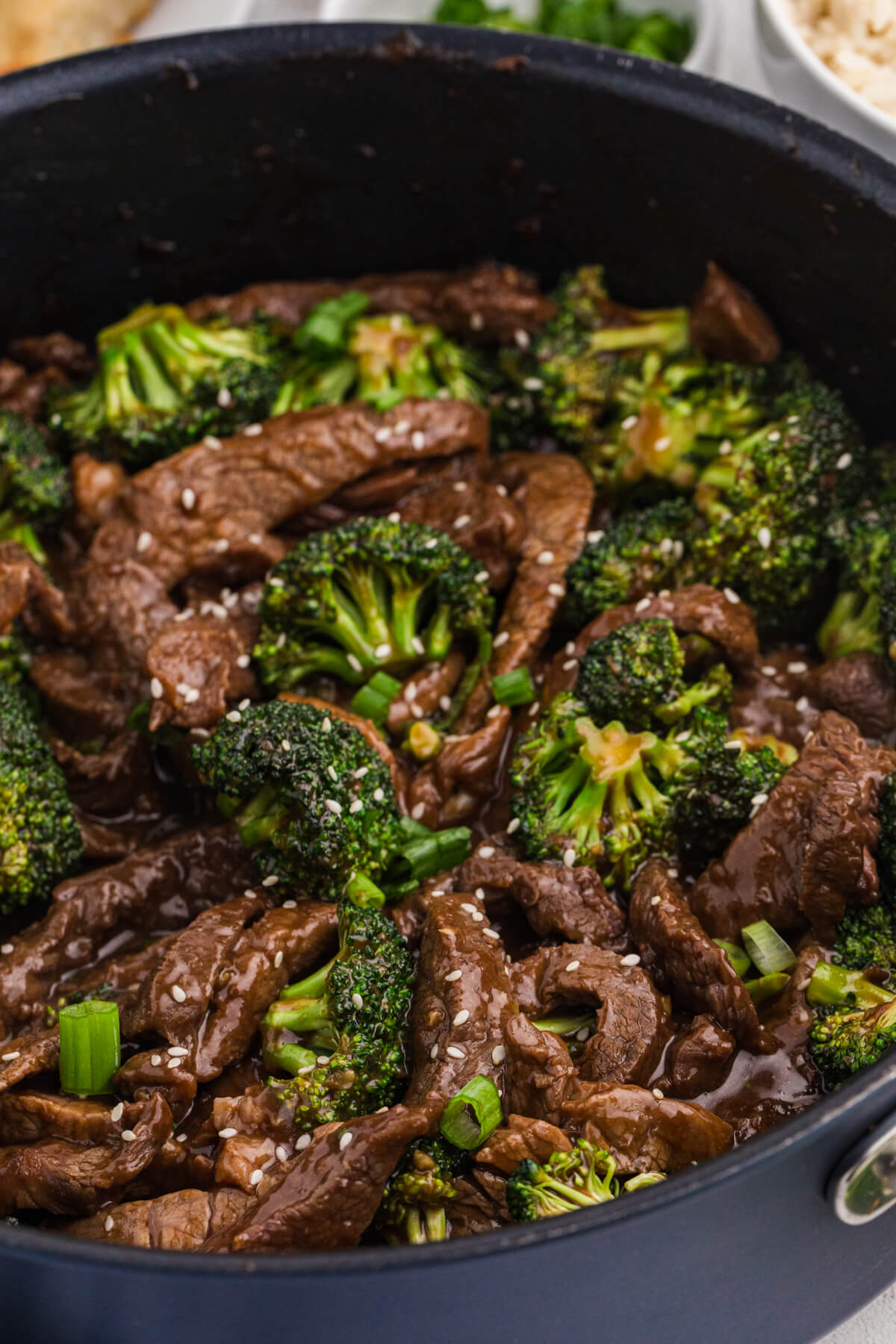 Close-up of beef and broccoli stir fry in a black pan, featuring tender sliced beef, bright green broccoli florets, sesame seeds, and a rich savory sauce, garnished with chopped green onions.
