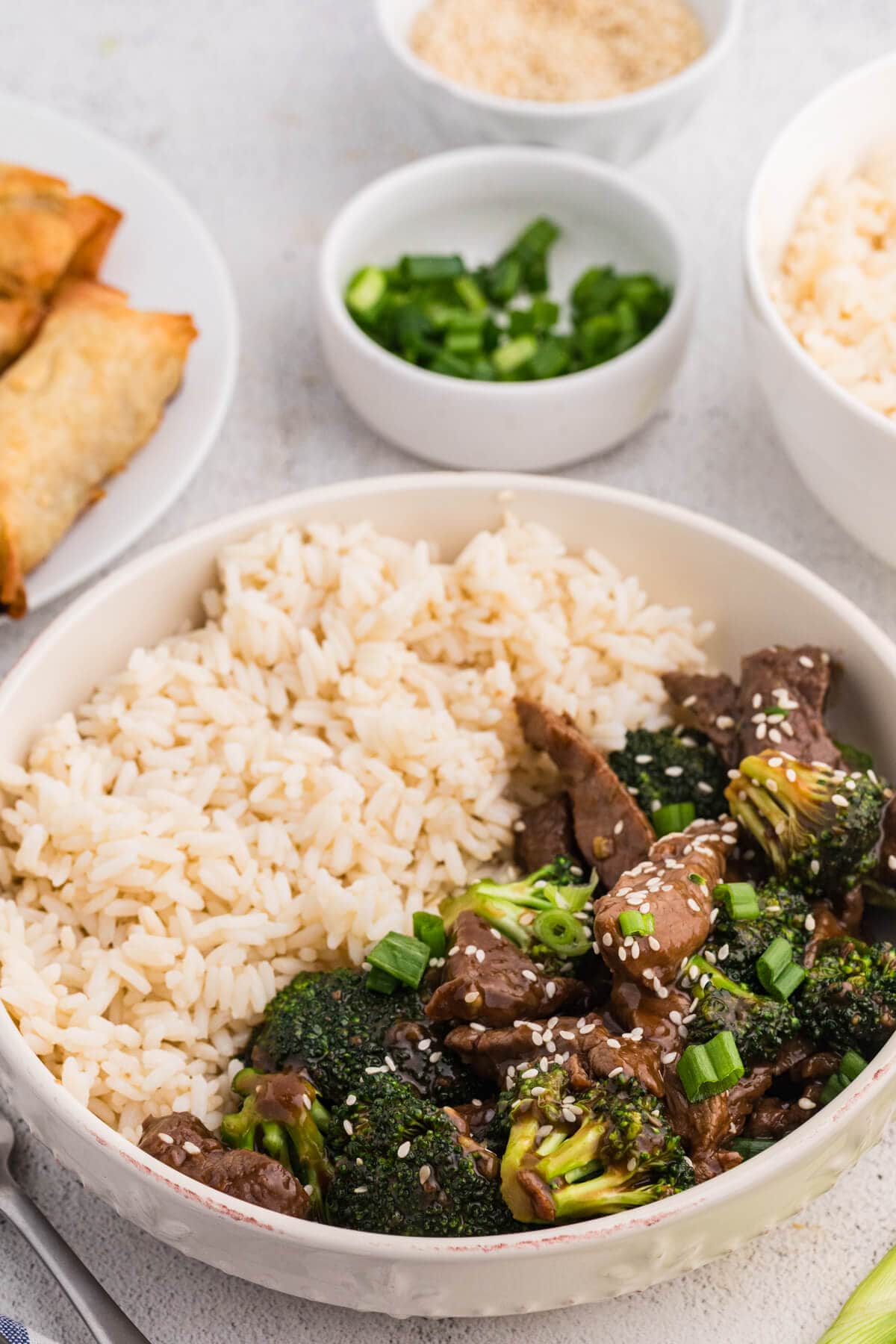Bowl of beef and broccoli stir fry topped with sesame seeds and green onions served with white rice, with spring rolls and small bowls of scallions in the background on a neutral kitchen surface.