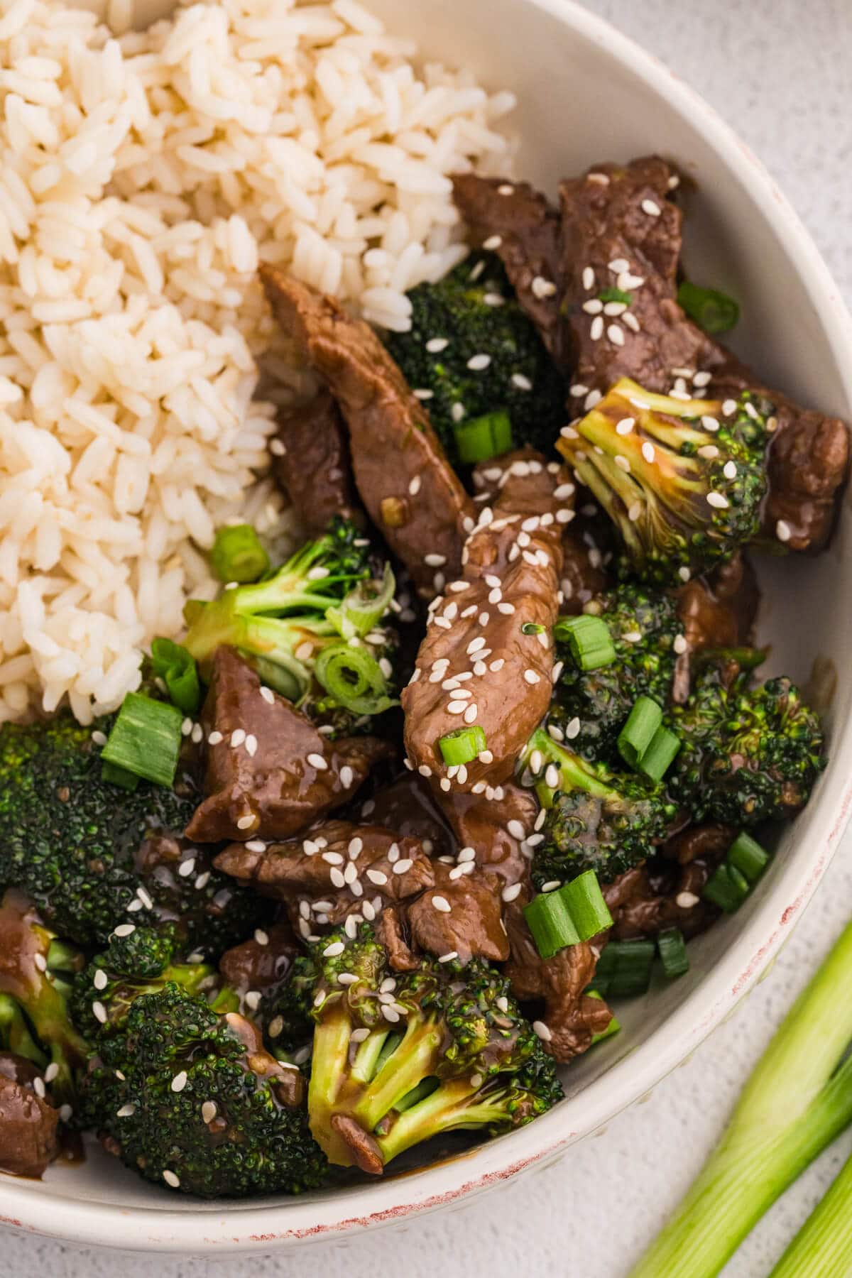 Detailed close-up of beef and broccoli stir fry with glossy sauce, sesame seeds, and chopped green onions next to fluffy white rice in a ceramic bowl.