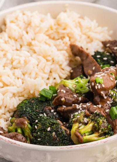 Serving bowl with beef and broccoli stir fry and steamed white rice, garnished with sesame seeds and green onions, with spring rolls and side dishes partially visible in the background.